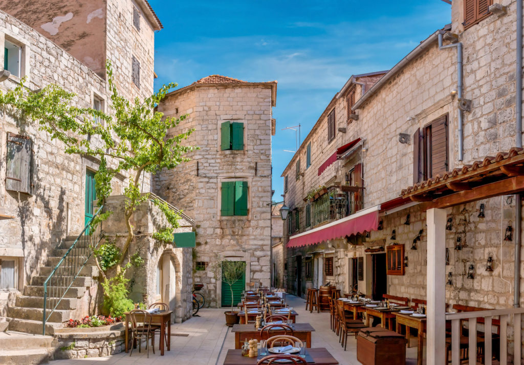 Street view of a charming outdoor tourist restaurant, open and prepared for customers, with rustic wooden furniture and surrounded by old stone buildings, in Stari Grad, Hvar Island, Croatia.