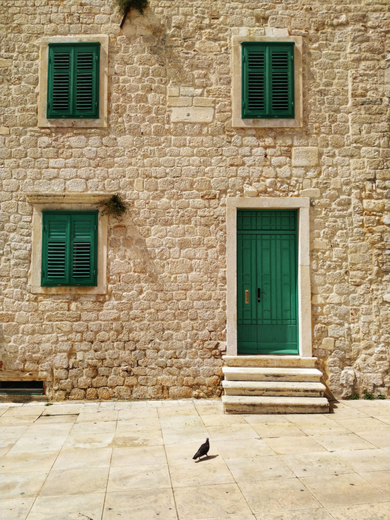 Green door, three shuttered windows and lonely dove in the yard of the house built of shell-rock stones
