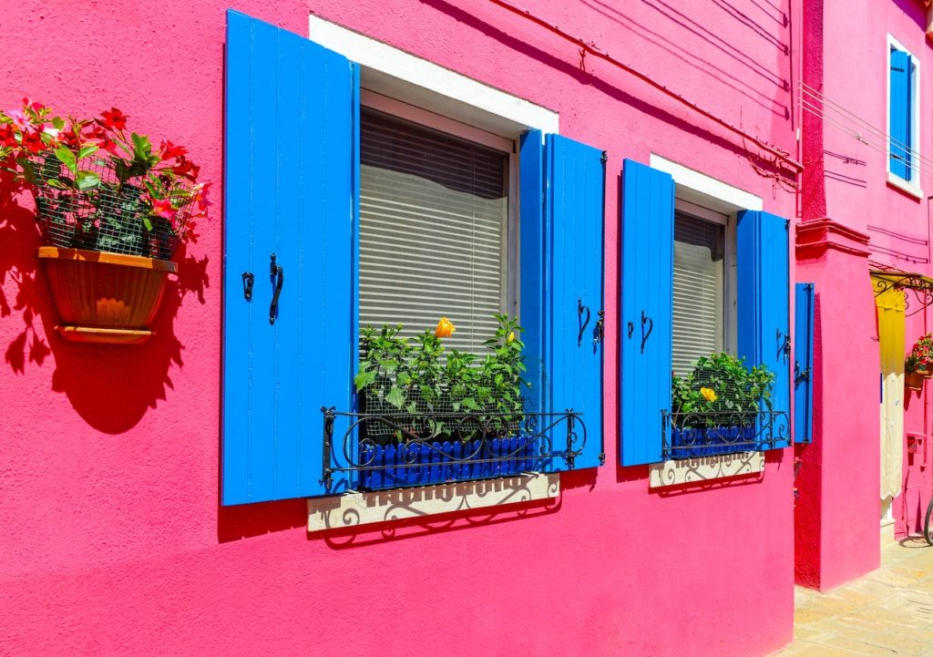 Flower pots decorate on the walls and blue windows of the pink house. Colorful architecture in Burano Island, Venice, Italy